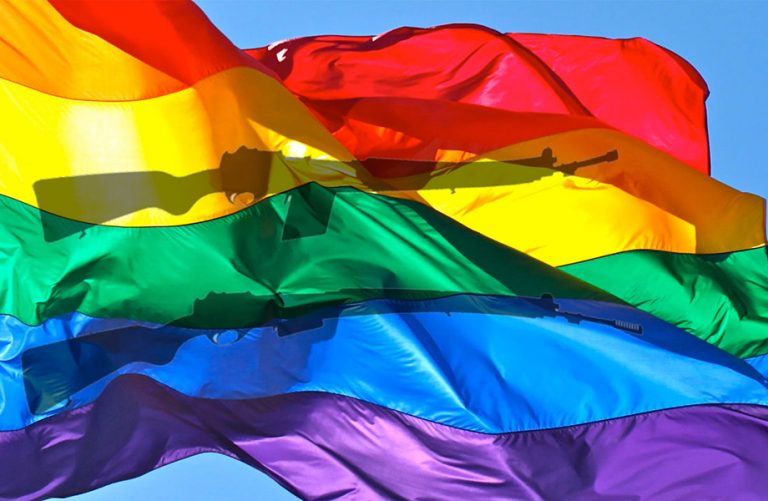 A rainbow flag flutters in the wind with a shadow of guns behind it.