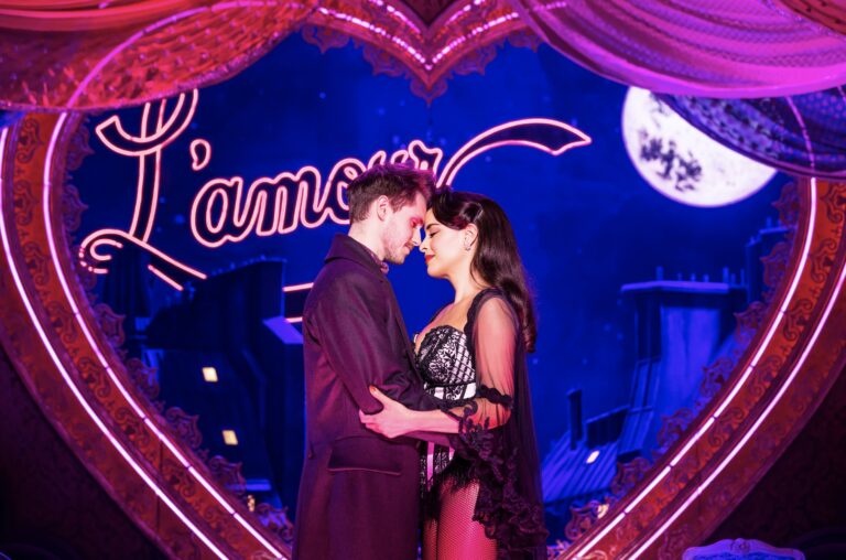 John Cardoza as Christian and Courtney Reed as Satine in the North American Tour of ‘Moulin Rouge! The Musical.’ The two actors are in a loving embrace as they stand in front of a lit up heart that says, 