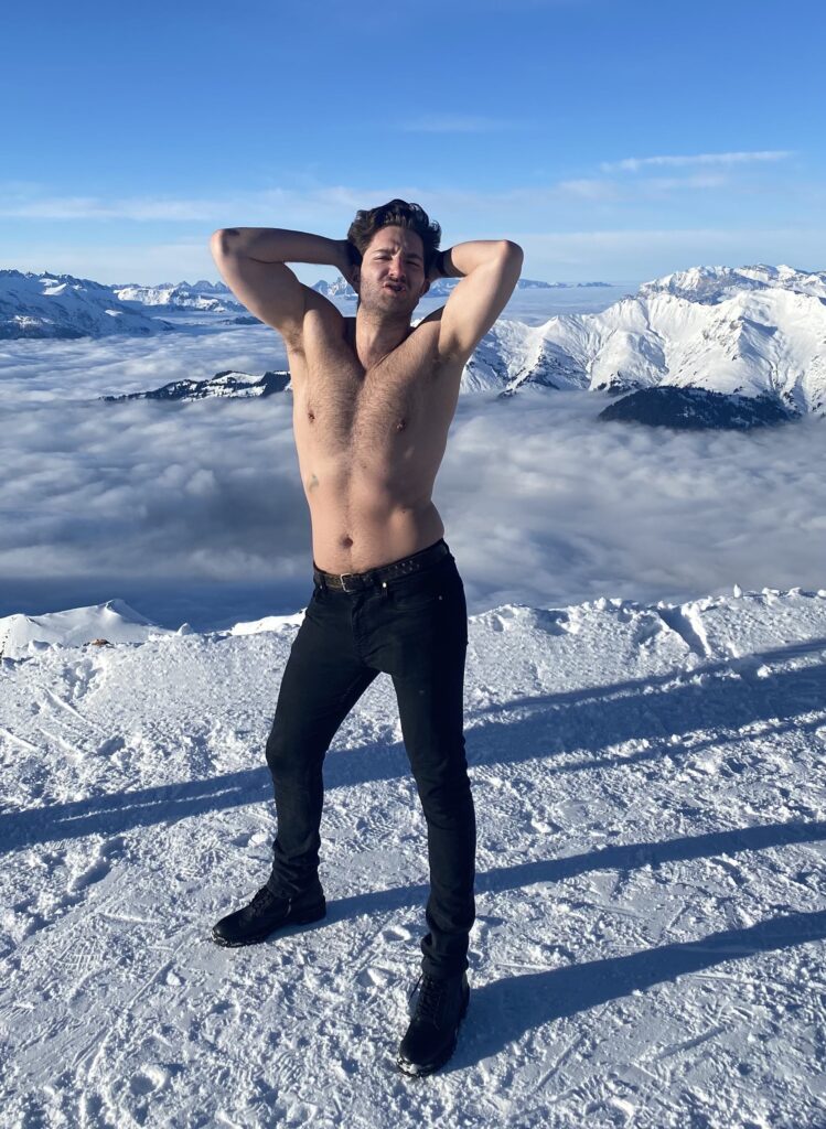 Zachary Zane stands shirtless while stretching his arms above the head while standing in the snow.