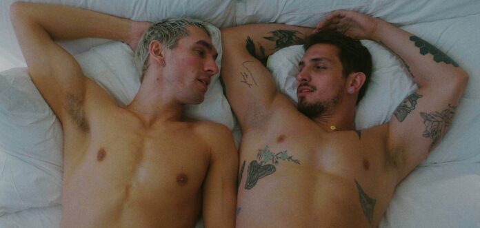 From left, Jack Stratton-Smith as Randy and Manuel Kornisiuk as Hunter in ‘The Winner Takes It All.’ The two men lay shirtless in bed together.