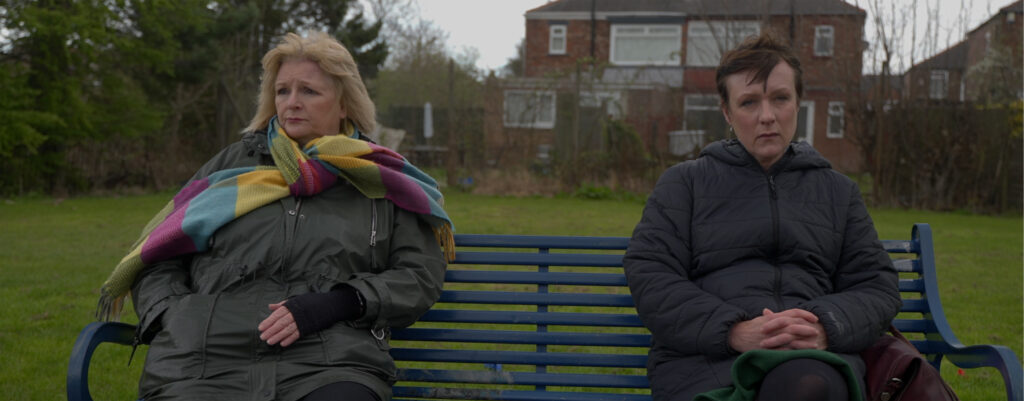 From left, Alex Hall as Amanda and Lucy Crawford as Cheryl in ‘Parallel Lies.’ The two are sitting on a bench looking off into the distance with despondent looks on their faces.