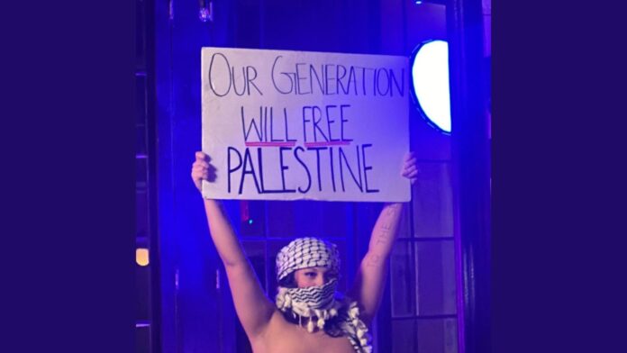 Leila Delicious, a burlesque artist, stands mostly nude during a recent performance at an unnamed venue, holding a sign that reads, “Our generation will free Palestine.” Her face and hair are covered by a scarf.