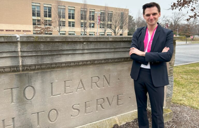 Jason Landau Goodman wears a dark suit and pink scarf. They are light-skinned with short, dark hair. They stand with arms crossed in front of Lower Merion High School in Ardmore.