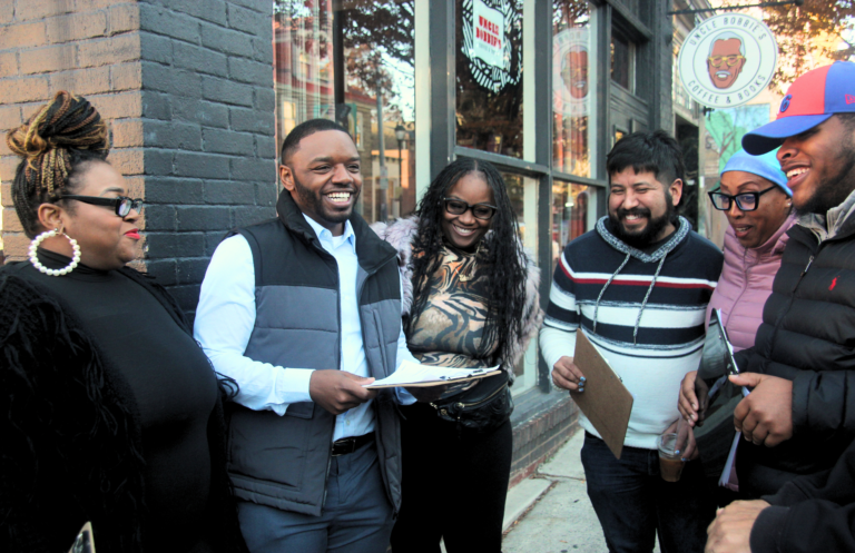 Andre D. Carroll stands among a group of people who are all people of color out front of a local coffee shop. They smile and laugh together, holding clip boards.