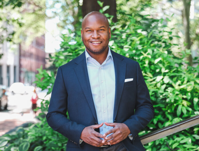 Michael Newmuis, a Black man, stands outdoors in front of greenery. He smiles his hands in front of his body, wearing a navy-colored suit and lighter blue button-down shirt.