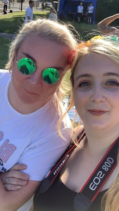Elizabeth is on the right and appears to have taken the photo, which is a selfie. They wear a black top and a camera strap around their neck. Shea is on the left with sunglasses and a white tee shirt. They are both light skinned with blonde hair.