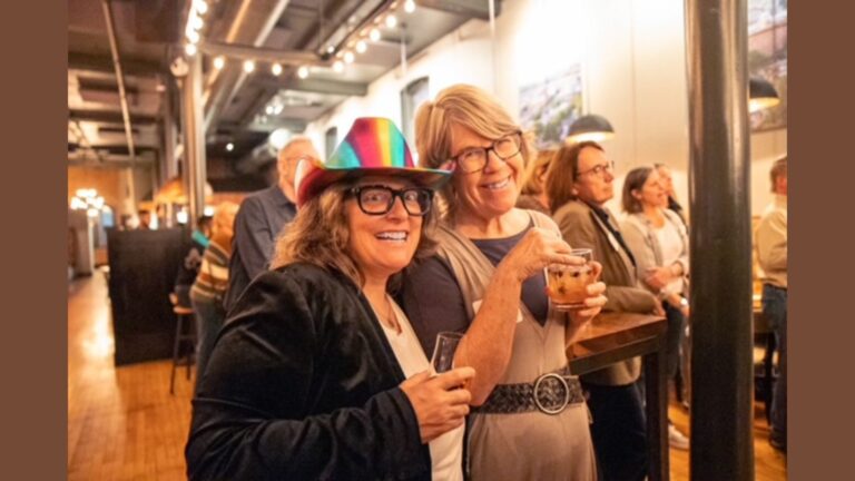 Suzette Mullen (who is light skinned with gray, chin-length hair) stands with her wife, Wendy (who is shorter than her and also light skinned with longer hair). Wendy wears a colorful cowboy hat and blazer. The couple holds their drinks amongst a crowd at an event.
