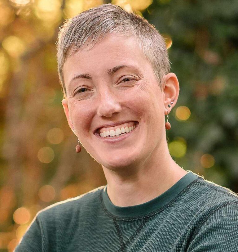 Alicia is a white person with short, gray hair that sits close to their head. They are seen smiling in front of a blurred outdoor background for a traditional headshot. They wear a deep green shirt and beaded earrings.
