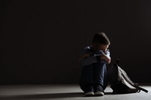 Upset boy with backpack sitting in dark room. Bullying concept