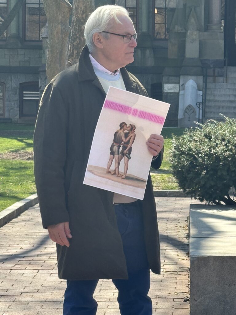 Jonathan Katz — a white man with white hair and glass — holds a sign that reads, “Queerness is historical." His sign displays an image of queer love. He wears a jacket and jeans.