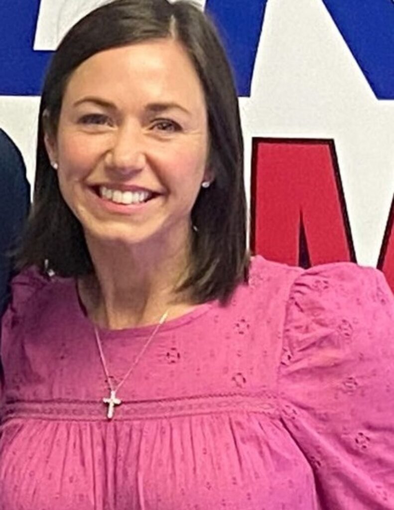 Katie Britt smiles and is wearing a pink shirt and necklace.