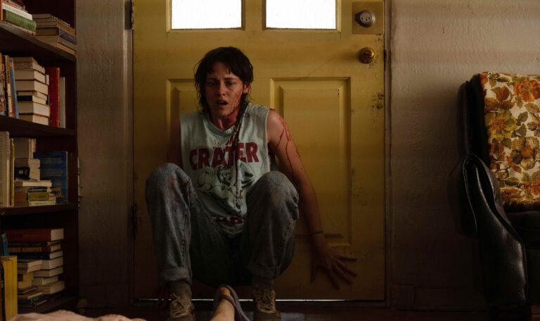 Kristen stewart sits in front of a door while covered in blood in a scene from 'Love Lies Bleeding.'