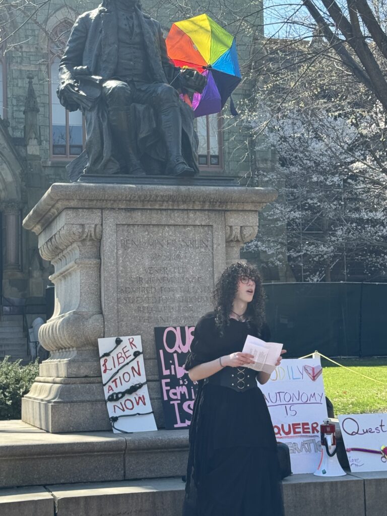 Lily Brenner wears a long, black dress. She stands in front of a statue of Benjamin Franklin and speaks to a crowd. The statue has protest signs and a rainbow umbrella has been placed in the hand of Benjamin Franklin to make it appear as though the statue is holding it.