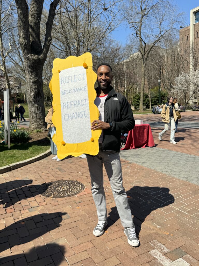 Malik Muhammad, a Black man wearing a casual sweatshirt and jeans with sneakers, stands while holding a sign that has a bold, yellow border. It reads, “Reflect resistance. Refract change.” He smiles.