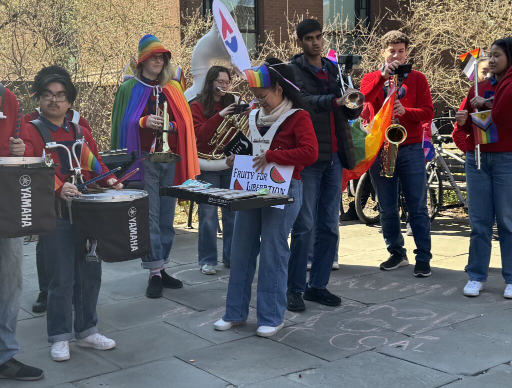 Members of University of Pennsylvania’s marching band — many wearing rainbow-colored clothing items in addition to their red Penn gear — perform on a paved courtyard out front of Penn's LGBT Center. They hold various instruments — including a tuba, drums, saxophone, tuba, xylophone — and some carry Pride flags.