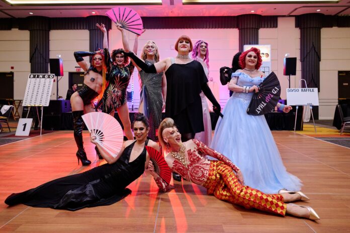 Two performers -- one in black and the other in red -- pose on the floor. Five others post standing above them. All wear colorful gowns and bejeweled costumes. Some hold decorative fans.