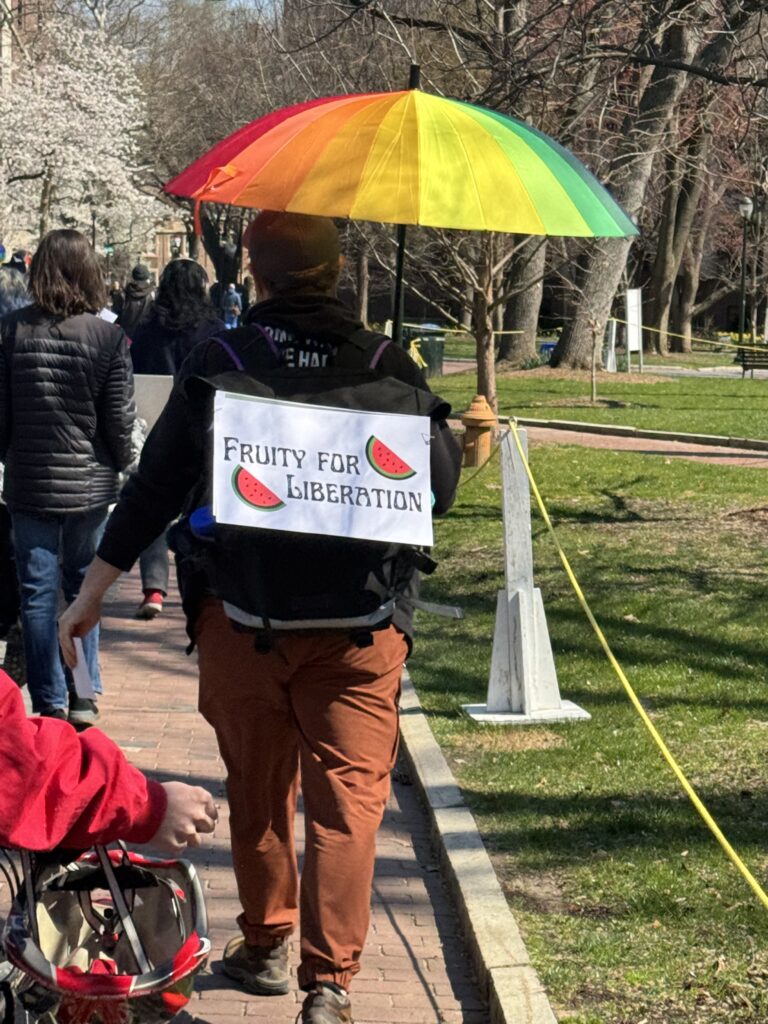 A protestor faces away from the camera. They march with a group that is not pictured, holding a rainbow umbrella. A sign hanging on their backpack reads, "Fruity for liberation."