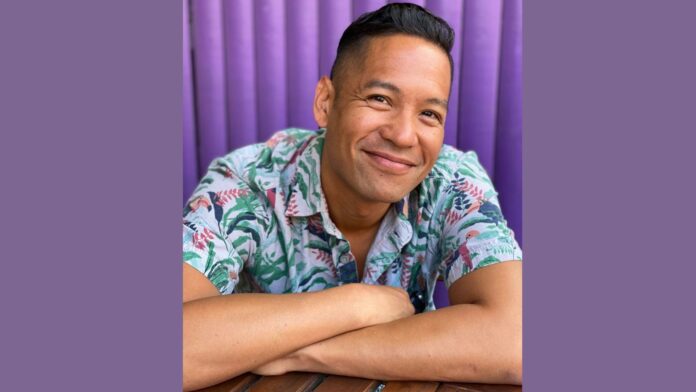 Justin Jain, an Asian American director, smiles with a closed mouth and soft eyes at the camera. His arms are crossed loosely on a table he leans against. He wears a colorful shirt.