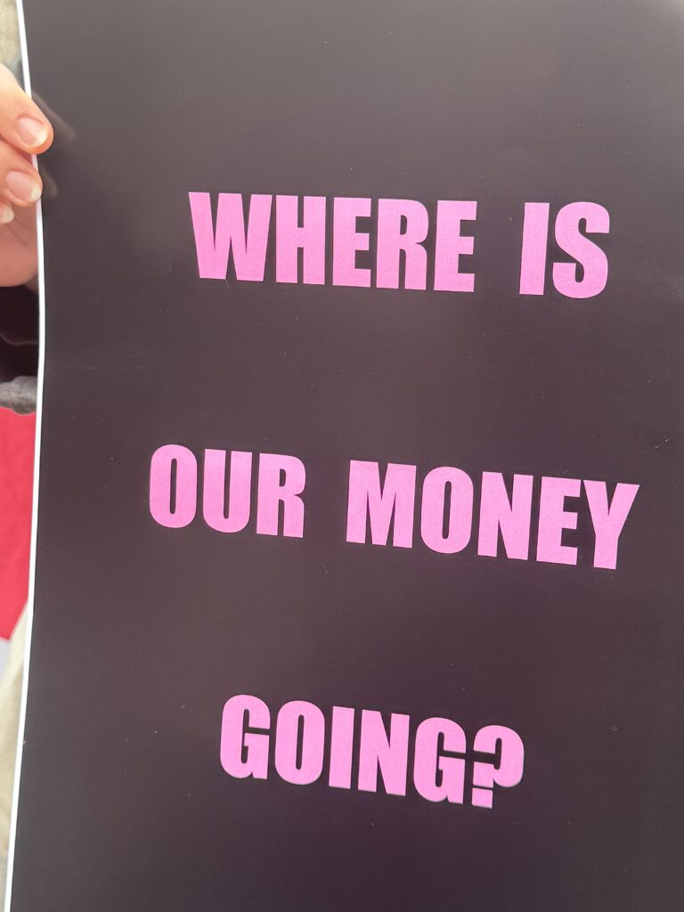 Fingertips can be seen holding the edge of a protest sign. It's a poster with a black background. In bright pink text, it reads in all caps, "Where is our money going?"