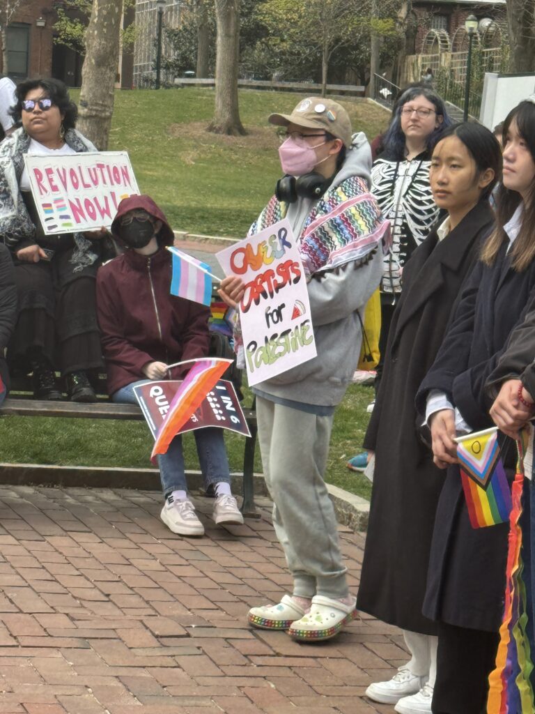 Activists gather to listen to speeches at Blanche Levy Park. One protester, who is wearing a colorful wrap around their shoulders, holds a sign that reads, “Queer artist for Palestine.” Some other signs and Pride flags are seen in the photo.