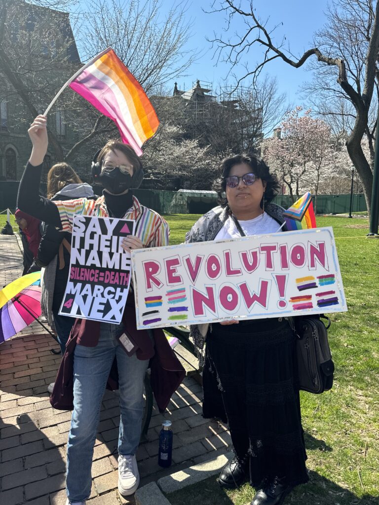 Two people stand and hold protest signs. One person waves a lesbian Pride flag. They hold a sign that addresses the death of Nex Benedict, reading "Silence = Death." The other holds a sign that displays various Pride flags and reads, "Revolution now!"