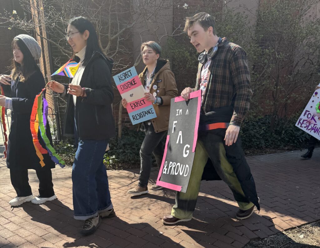 Protesters carry Pride flags, colorful ribbons, and signs as they march. One reads, “I’m a fag and proud.” The other says, “Respect existence or expect resistance.”