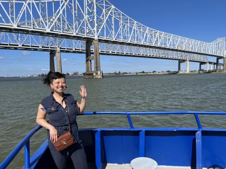 Diana Lu is on a ferry or boat. She waves to the camera on an industrial tour as part of the Greater Philadelphia Leadership Exchange. A bridge is behind her. She wears a denim, sleeveless jumpsuit. Her dark hair is in a ponytail and shows her bangs.