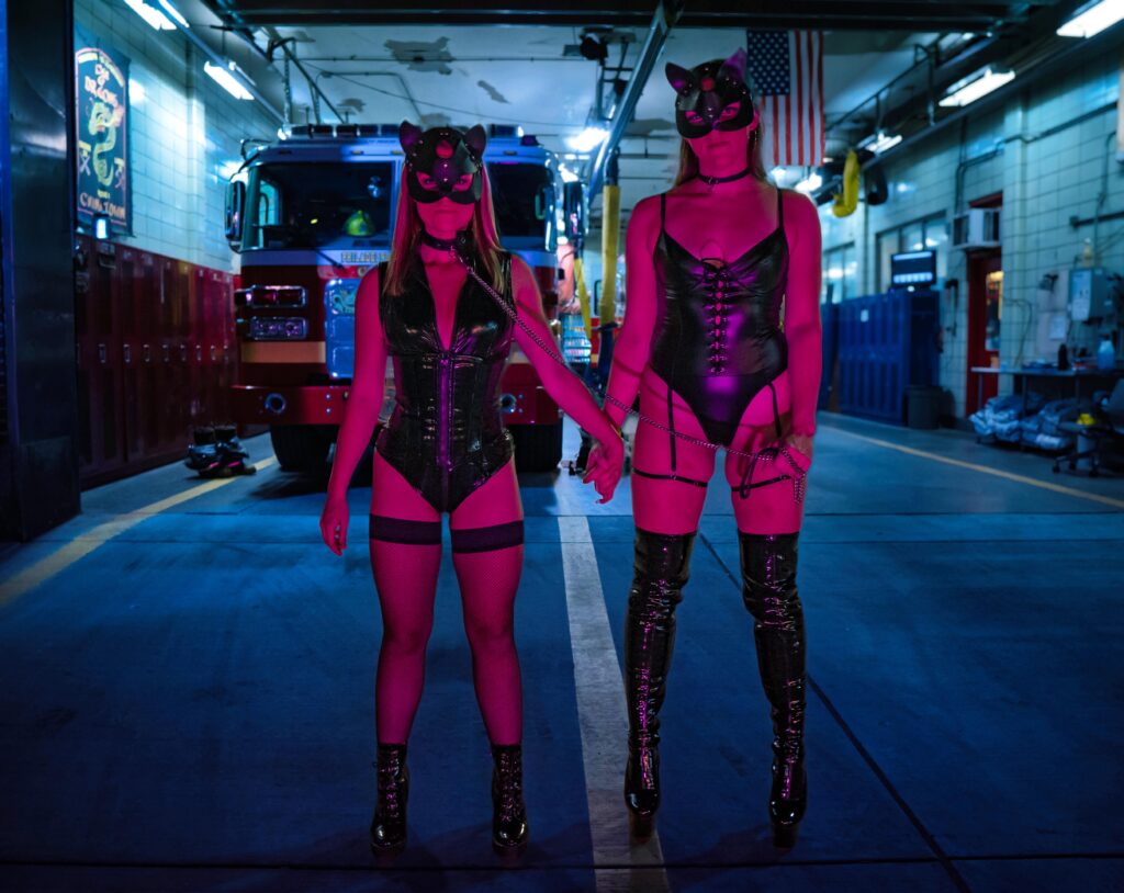 Two people dressed in leather bunny outfits pose in front of a fire house.