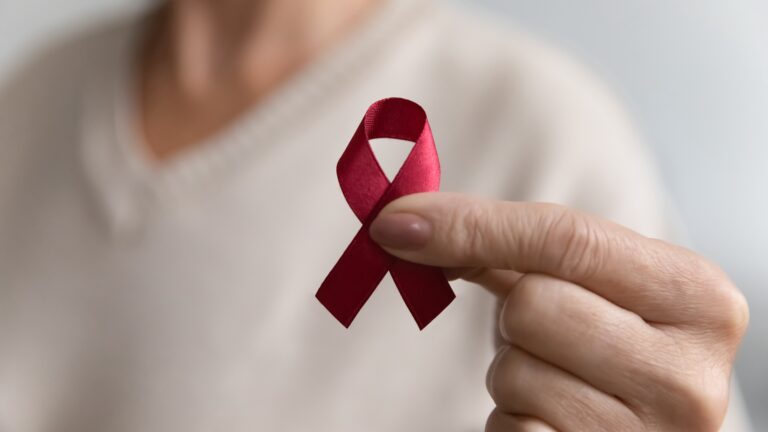 A light-skinned person holds a small red ribbon for HIV awareness in front of their chest. They wear a white sweater and have very pale pink nail polish on.