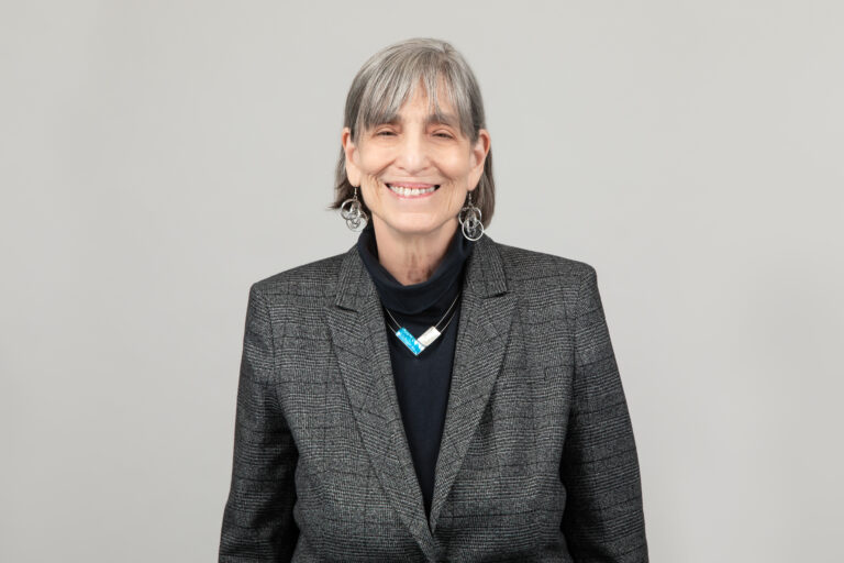 A white woman smiles at the camera. She has gray hair that is sits just above her shoulders and bangs. She wears a gray suit jacket and black shirt with a blue and white necklace near the collar.