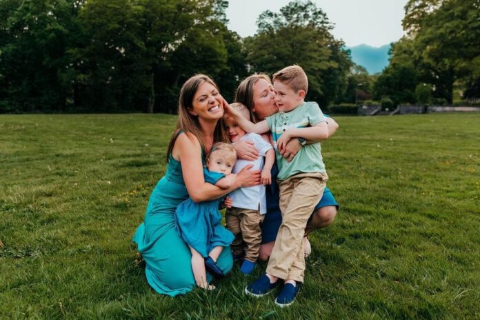 Two moms with three kids smile in the grass.