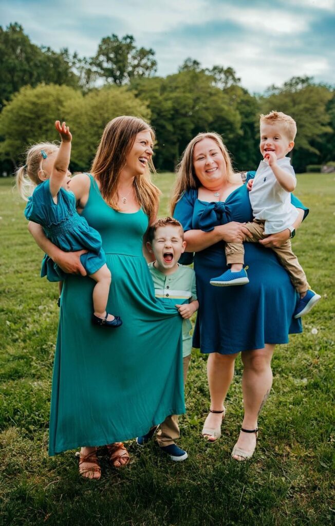 Two moms smile in the grass while embracing their three kids.