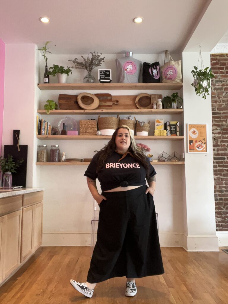 Julia Birnbaum wears a black t-shirt with the word "Brieyonce" on the front and a long black skirt. She has her hands on her hips as she poses in front of shelves