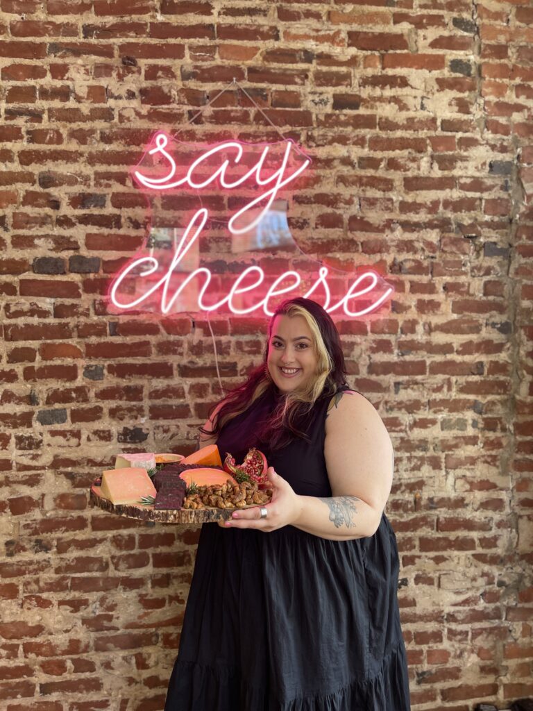 Julia Birnbaum holds a platter of cheeses in front of a light-up sign that says "say cheese"