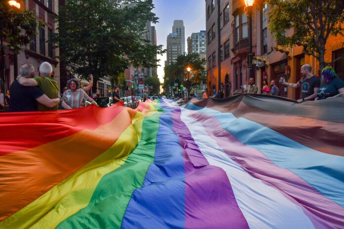 A massive Pride flag reaches in length so far the eye can't see its beginning and ends. It spans the width of the street. Two people embrace nearby while some people hold onto the flag from the sides of the road. Philadelphia buildings -- including Center City's Comcast building -- is visible in the background.