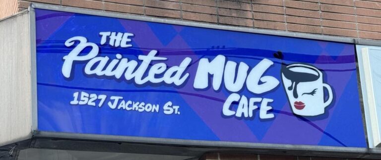 The sign that sits above the main entrance of The Painted Mug Cafe. It's a blue background with white text. The logo, a coffee mug with a face styled in make-up, is pictured.