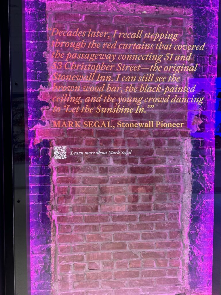 A sealed passageway that previously connected the building that currently houses the visitor’s center with the Stonewall Inn next door. On glass in front of the wall, a quote from Mark Segal reads, “Decades later, I recall stepping through the red curtains that covered the passageway connecting 51 and 53 Christopher Street — the original Stonewall Inn. I can still see the brown wood bar, the black-painted ceiling, the young crowd dancing to ‘Let the Sunshine In.’”