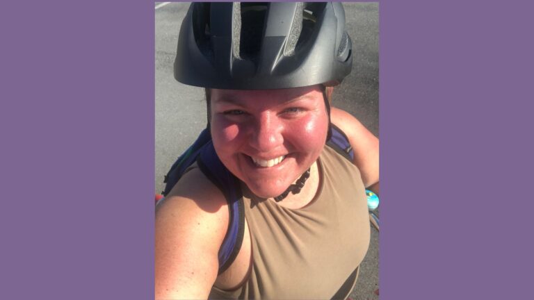 Amber Roadcap smiles at the camera for a selfie. She is a light-skinned person and wears a helmet and tank top. She sits atop her bike, which is not in the frame of the picture.