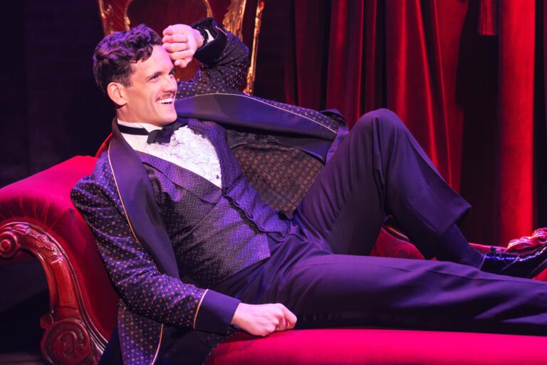 Stephen Mark Lukas as Nick Arnstein in the national tour of ‘Funny Girl.’ He is smiling while sitting on a couch with his hand on his forehead.