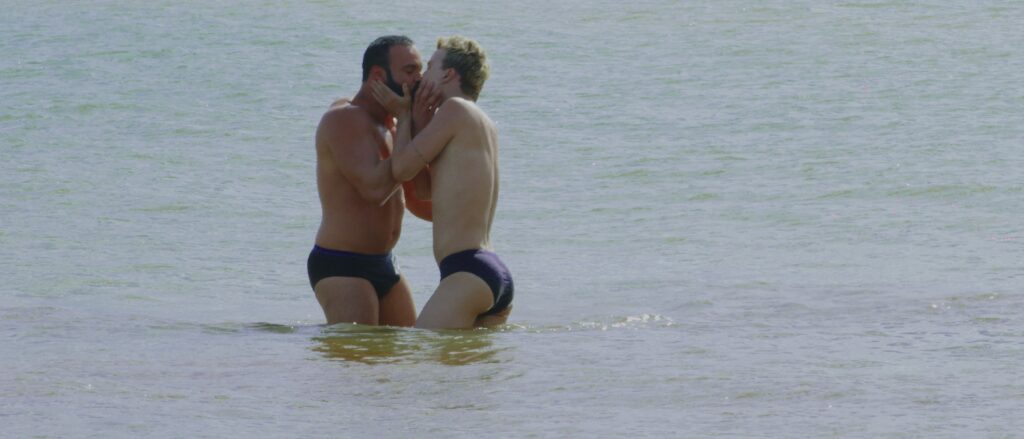 Two men in speedos kiss in the ocean.