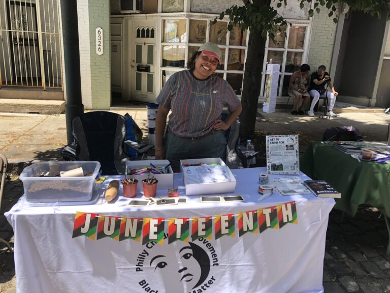 Tara Rose Brown smiles at the camera with her hand on her hip. She stands behind a table with various activities, planting materials, and information available at an event. The table displays a banner in celebration of Juneteenth.