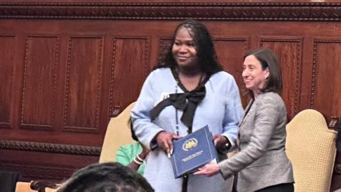 Tatyana Woodard stands next to Rue Landau. They smile toward a focal point off camera and together hold a blue folder that notes it is from the mayor's office.