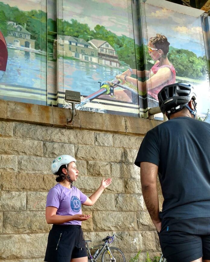 Rebecca Fisher -- pictured in a purple tee shirt, black shorts, and bike helmet -- gestures to a mural as she leads a tour. An attendee is pictured in front of her and off to the side, observing and listening.