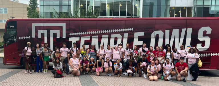 More than two dozen Temple students stand and kneel in front of a Temple bus.