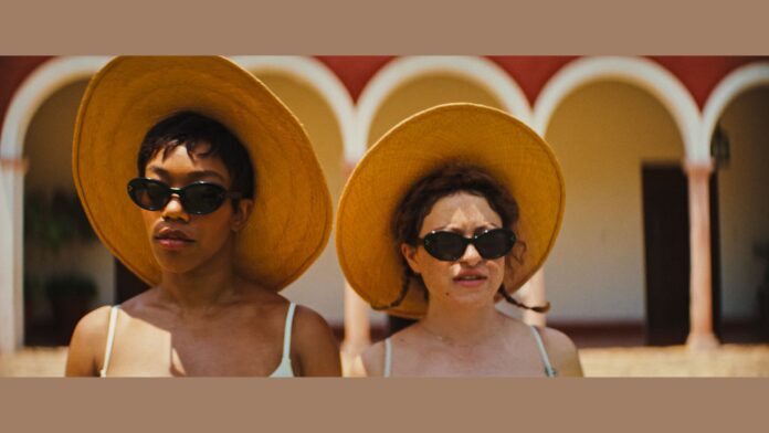 Two women are featured in this scene from the movie "Blink Twice." Both are wearing large, wide-brimmed yellow sun hats and dark sunglasses, giving them a stylish and somewhat mysterious appearance. The woman on the left has short, dark hair and wears a serious expression, while the woman on the right has lighter, curly hair with small braids, and a more neutral expression. They are dressed in light-colored, sleeveless outfits. The background features an outdoor setting with arches and a warm, sunlit ambiance, contributing to the summery and relaxed vibe of the scene.