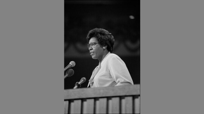 The image is a black-and-white photograph of Barbara Jordan, an influential American politician and civil rights leader, standing at a podium with two microphones. She is wearing glasses and a light-colored blouse with a scarf or bow tied at the neck. Barbara Jordan appears to be delivering a speech, with a focused and serious expression. The background is out of focus, highlighting her presence and the significance of the moment.