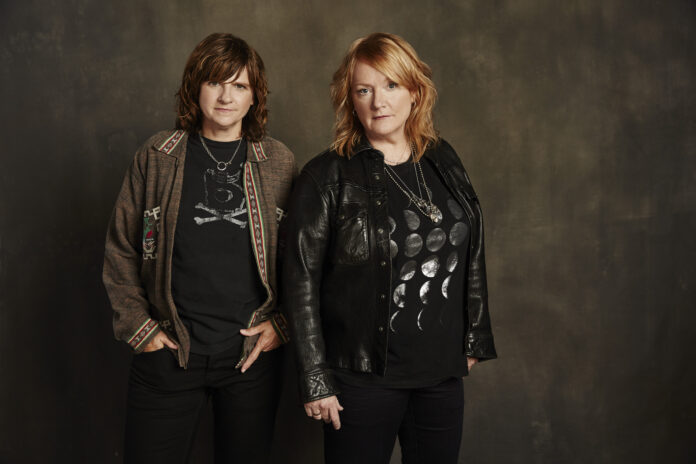 From left, Amy Ray and Emily Saliers stand while wearing jackets and looking confidently into the camera.