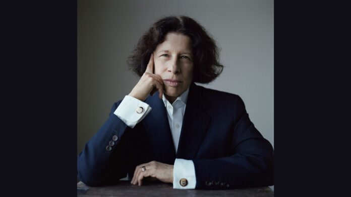 Fran Lebowitz is seated and dressed in a dark suit jacket and white collared shirt, her hand resting on her face with her index finger touching her temple. Her expression is serious and contemplative, with her dark, slightly wavy hair framing her face. The background is a muted gray, emphasizing her thoughtful demeanor.