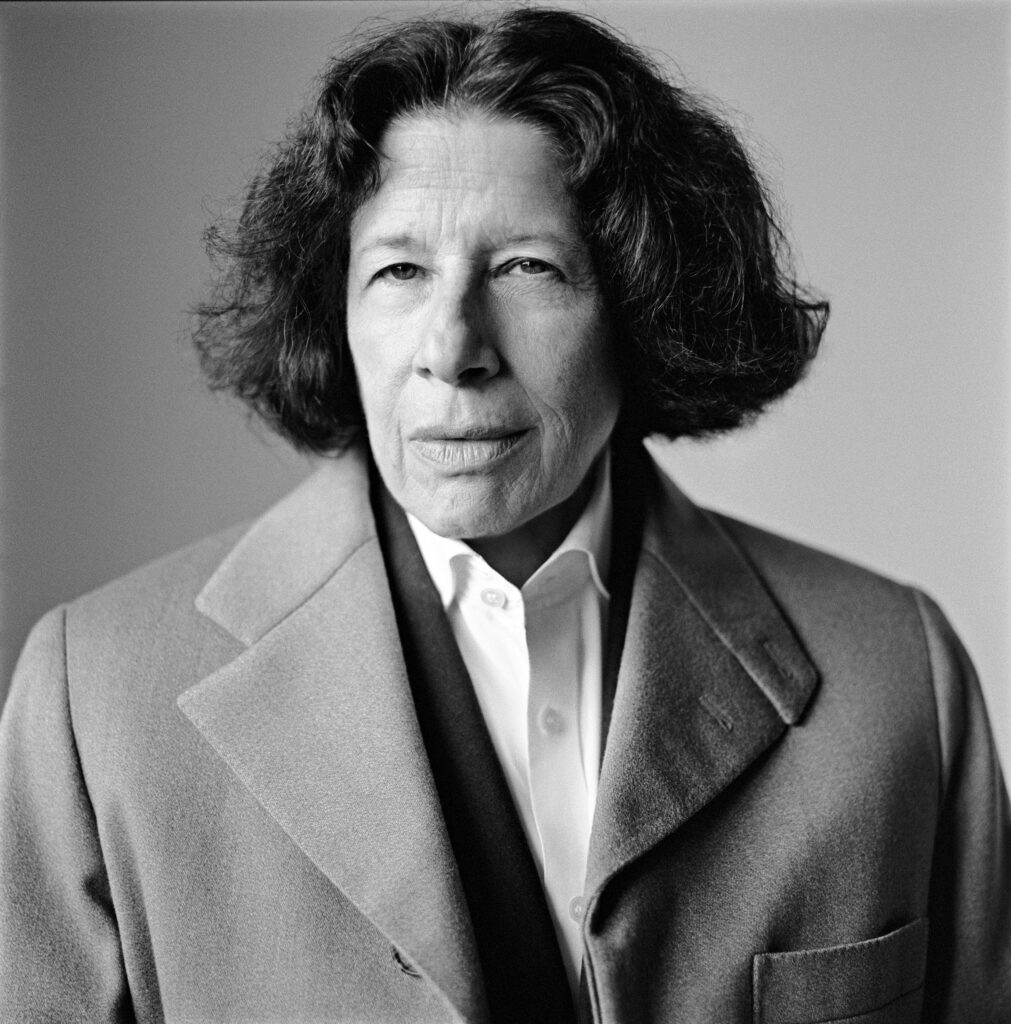 Fran Lebowitz stands facing the camera, wearing a light-colored overcoat over a white collared shirt. Her expression is intense and focused, with her lips slightly pursed. The black and white format highlights the texture of her hair and the lines on her face, giving the portrait a classic and timeless quality. The background is plain, drawing attention to her sharp gaze.