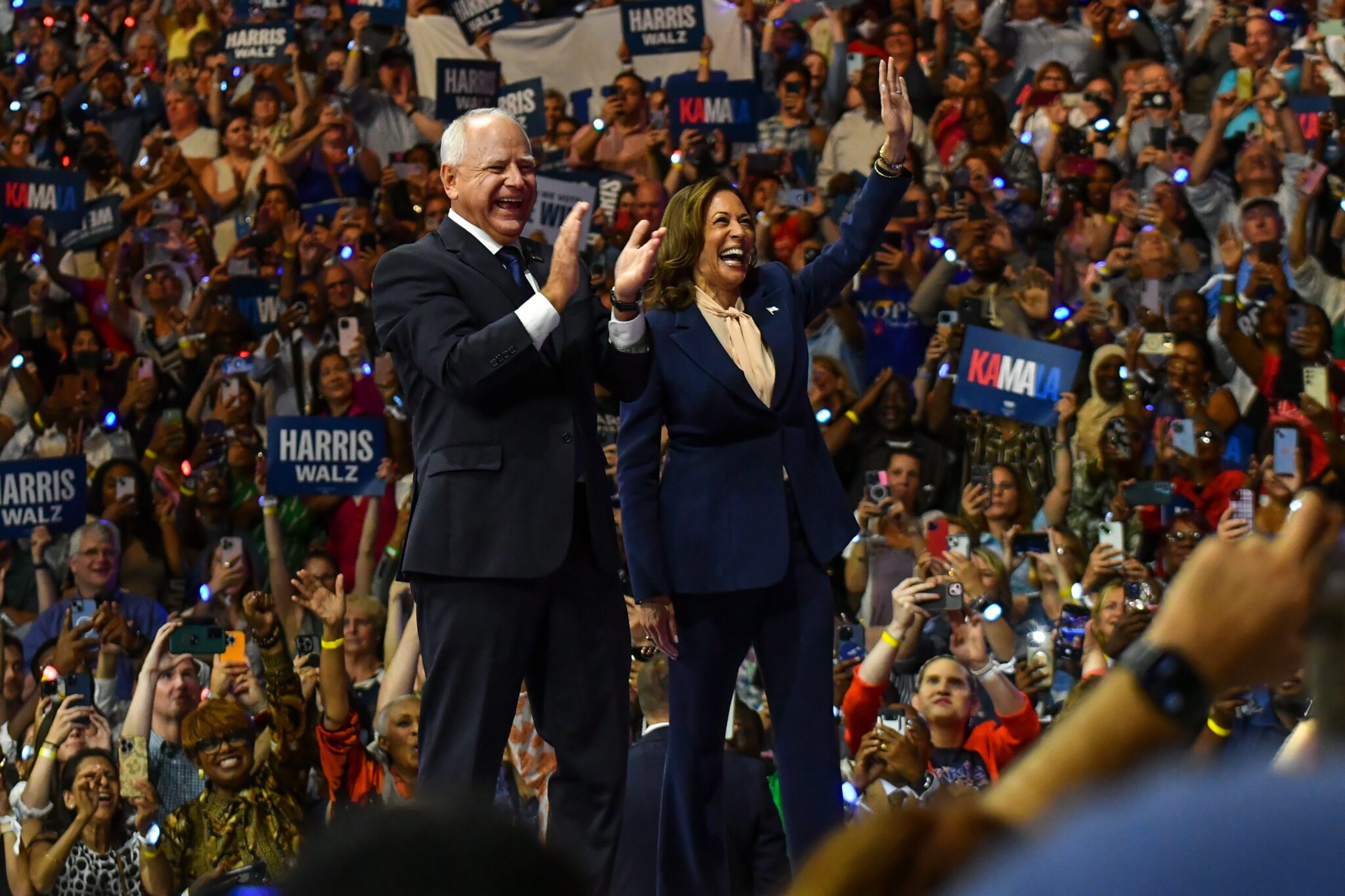 Kamala Harris and Tim Walz wave and clap in front of a large crowd.